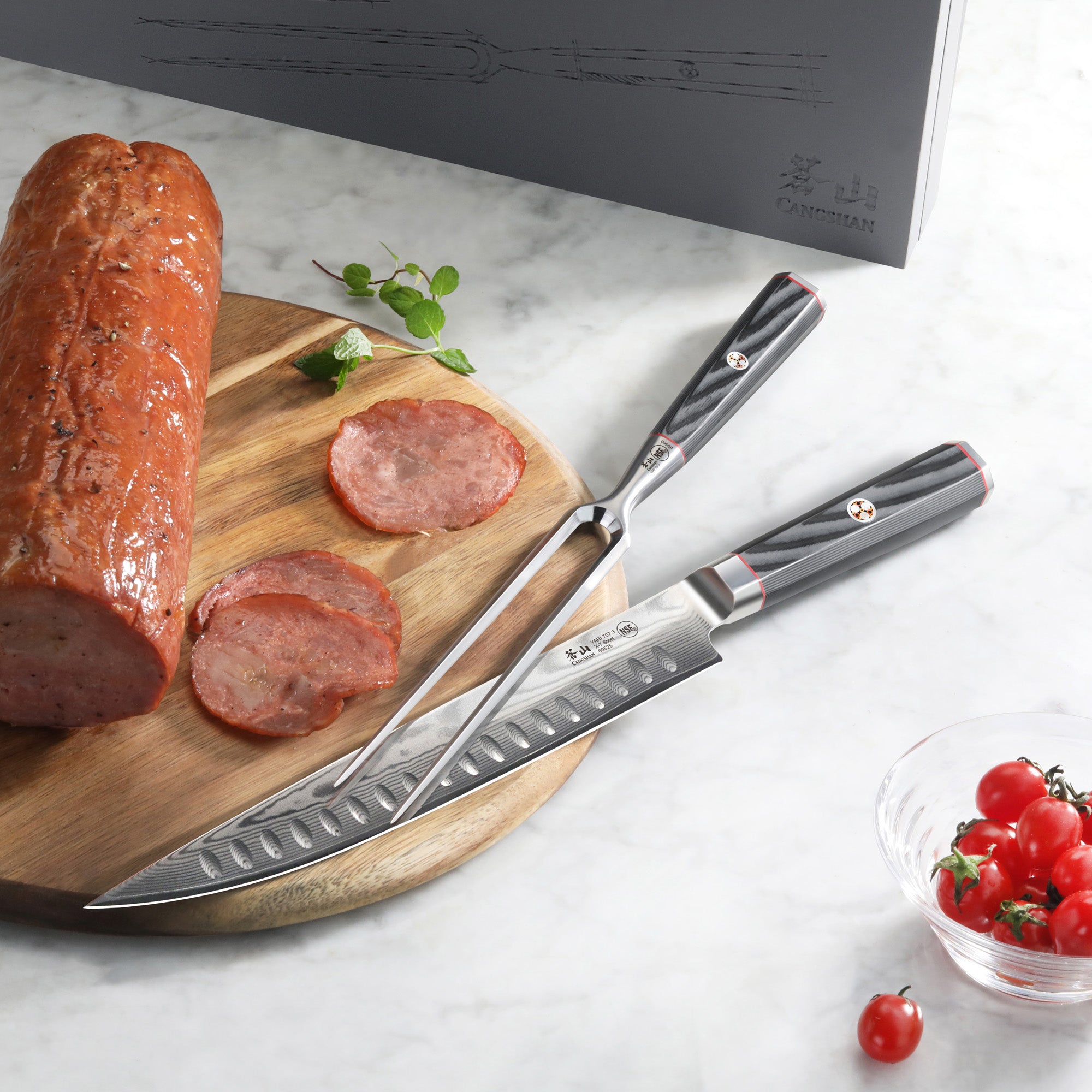 Two-piece carving set with black handles on a marble surface, next to sliced meat on a wooden board and a bowl of cherry toma