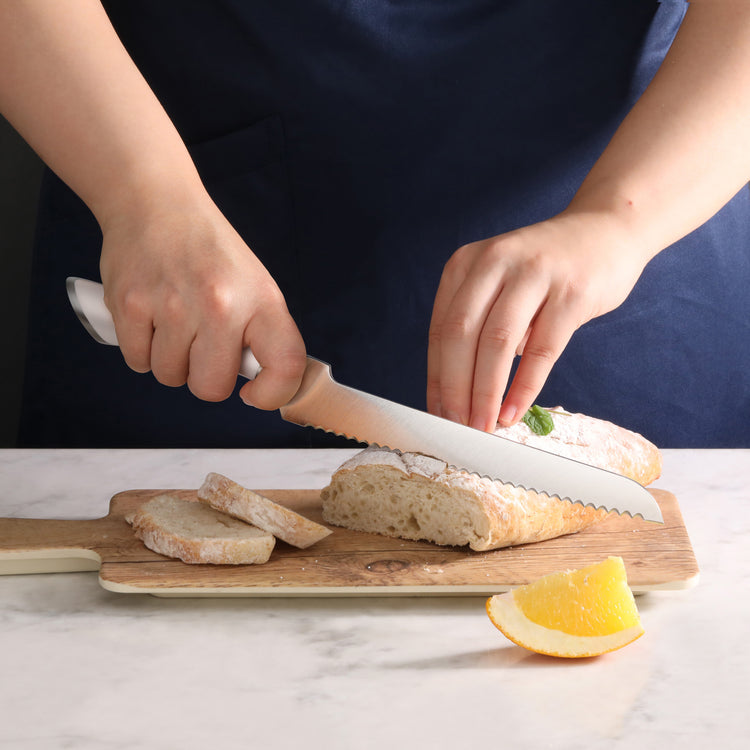 Hands slicing a loaf of bread with an 8-inch serrated bread knife on a wooden cutting board.