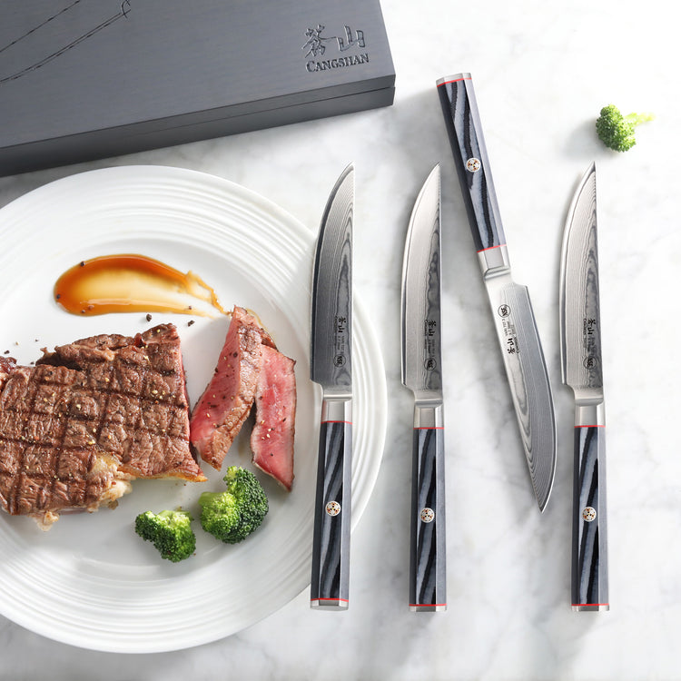 Four steak knives with patterned blades and dark handles arranged beside a plate with grilled steak slices and broccoli on a