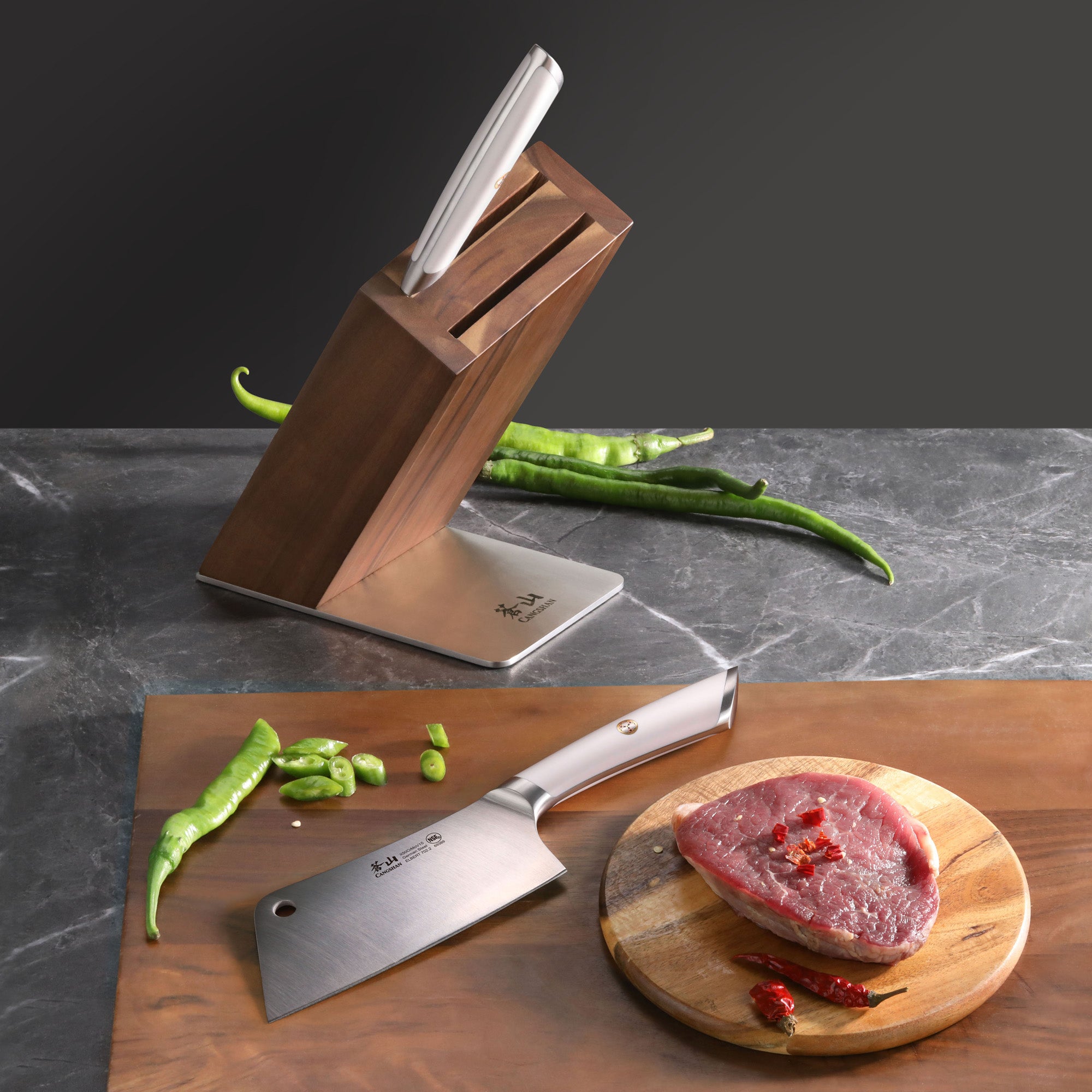 Knife block set with a cleaver knife on a cutting board next to a piece of raw meat and green chili peppers.