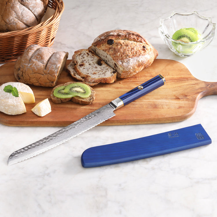 9-inch serrated bread knife with blue handle and sheath on wooden board with sliced bread, cheese, and kiwi.