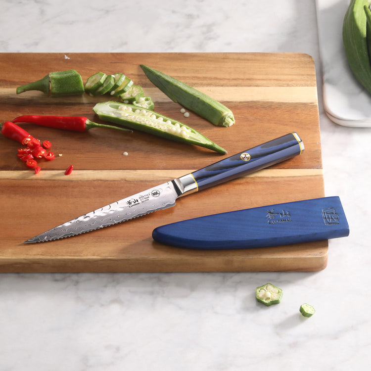 Serrated utility knife with blue handle and sheath on wooden cutting board with sliced okra and red chili peppers.