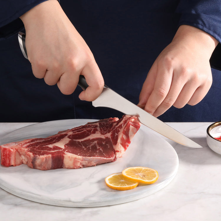 Person holding a boning knife cutting a raw steak on a round marble board with lemon slices nearby.
