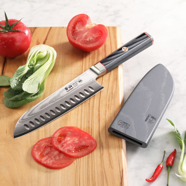 7-inch Santoku knife with patterned Damascus steel blade and black handle, next to gray sheath and sliced tomatoes on wooden