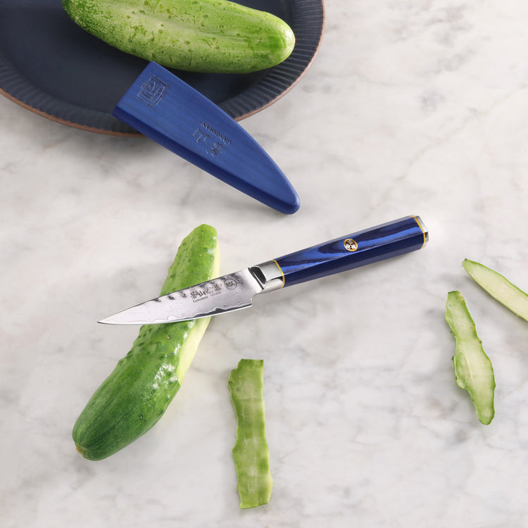 Paring knife with blue handle and sheath next to a partially peeled cucumber on a marble surface.