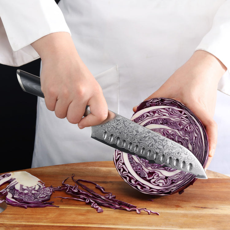 Hand holding a patterned 7-inch Santoku knife slicing a halved purple cabbage on a wooden cutting board.