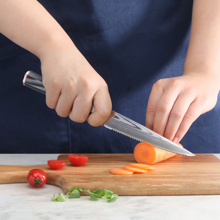 Hand holding serrated knife slicing a carrot on a wooden cutting board with cherry tomatoes and basil nearby.