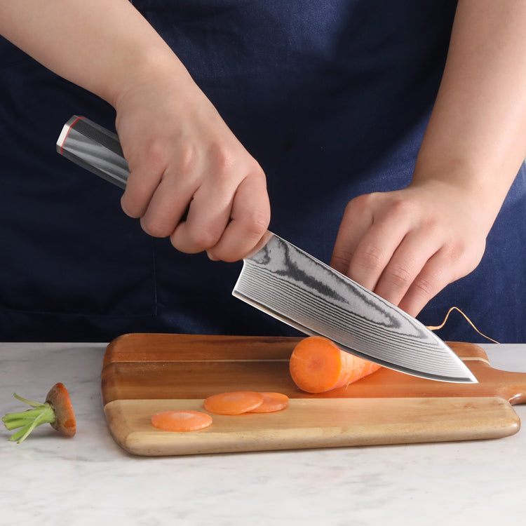 Hand holding a Damascus steel knife slicing a carrot on a wooden cutting board.