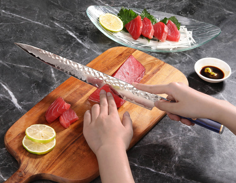Hands slicing raw tuna on a wooden board with a blue-handled sashimi knife, plate of tuna sashimi and lemon slices nearby.