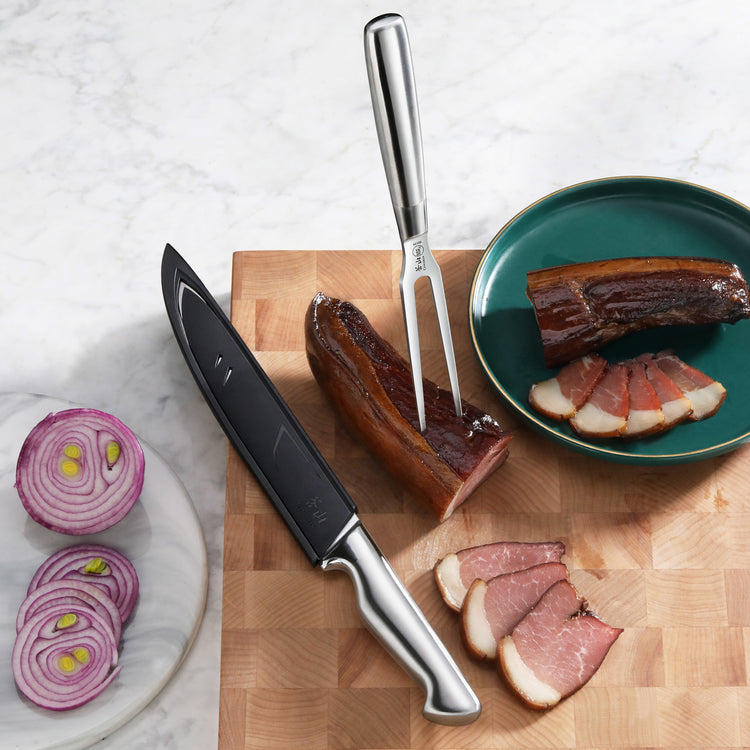 Carving knife and fork on wooden board with sliced smoked meat, green plate, and sliced red onion on marble surface.