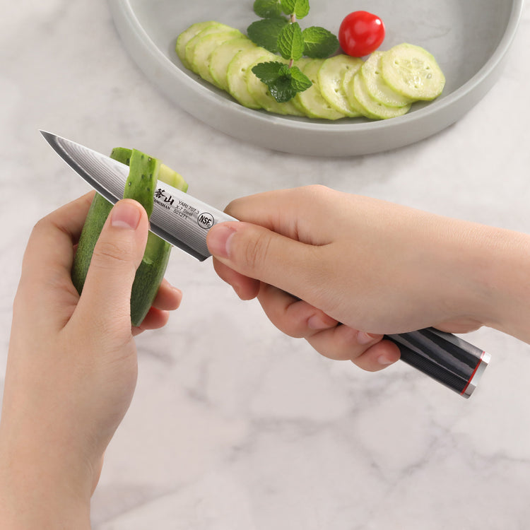 Hands peeling a cucumber with a small paring knife featuring a black handle and Damascus steel blade.