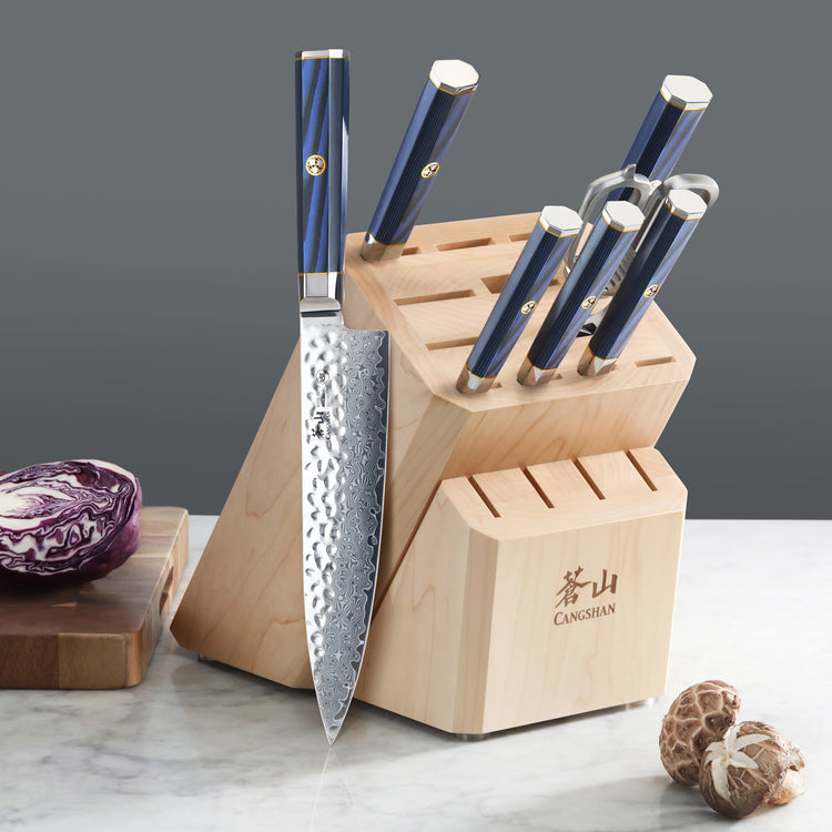 Wooden knife block with six blue-handled knives inserted and two empty slots, one large knife resting on the block.