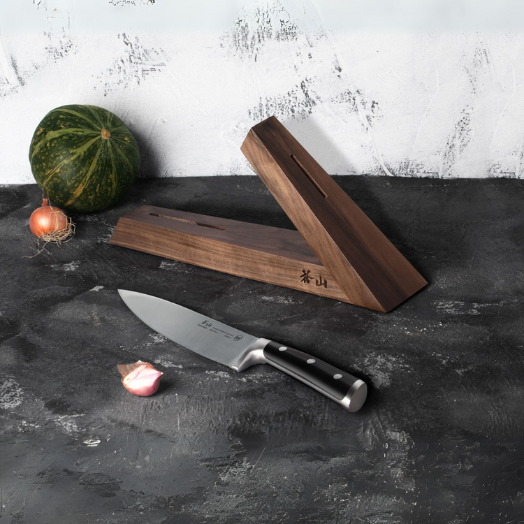 Walnut wood triangular knife block with a chef's knife and small vegetables on a dark surface.