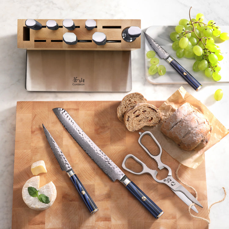 Set of three Damascus steel knives with blue handles on a wooden board, accompanied by bread, cheese, and grapes.