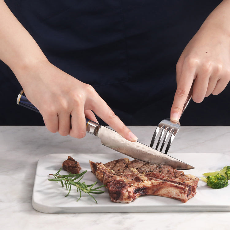 Hands using a steak knife and fork to cut a cooked T-bone steak on a white cutting board with herbs and broccoli.