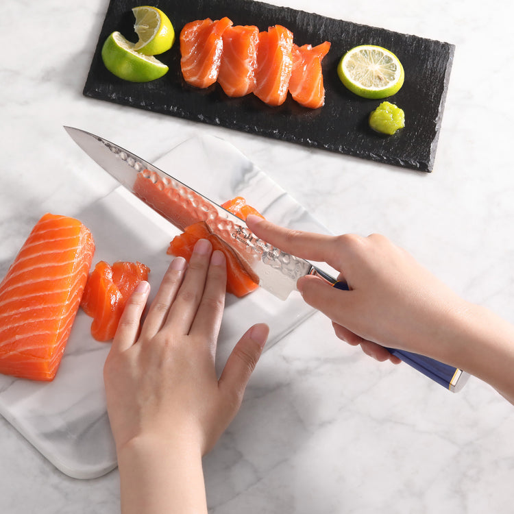 Hands slicing fresh salmon sashimi with a blue-handled knife on a white cutting board, with lemon and wasabi nearby.