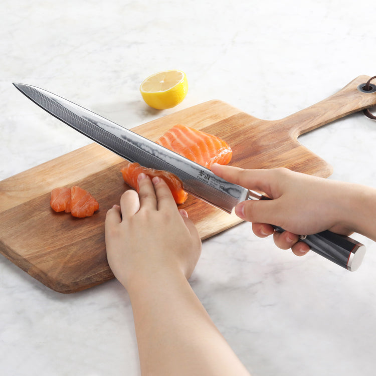 Hand holding a sashimi knife slicing raw salmon on a wooden cutting board with half a lemon in the background.