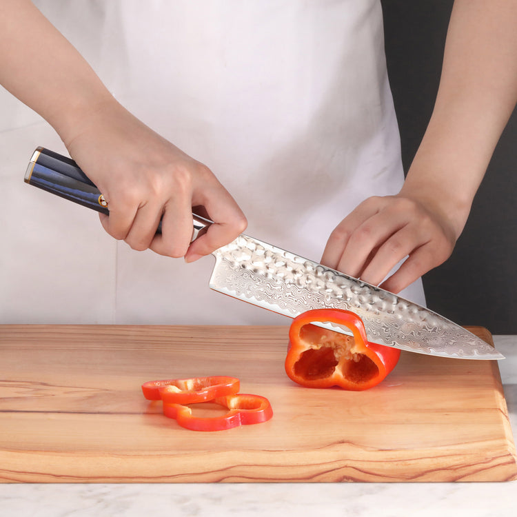 Person slicing a red bell pepper on a wooden cutting board with a hammered steel knife.