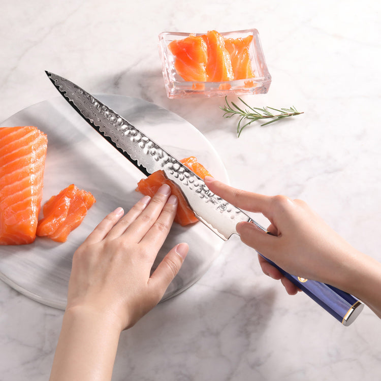 Hands slicing raw salmon on a white plate using a 10-inch sashimi knife with a blue handle.