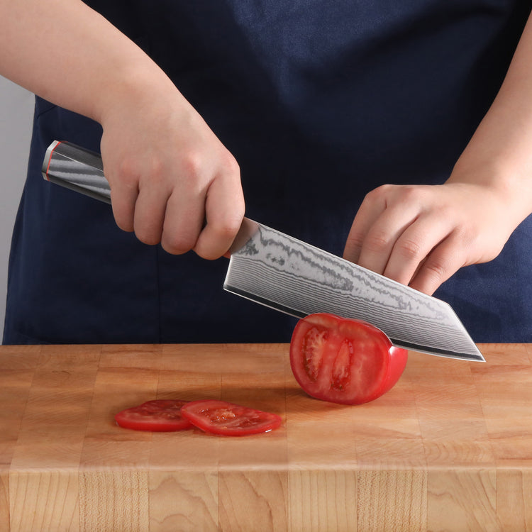 Person slicing a tomato on a wooden cutting board with a patterned blade Kiritsuke knife.