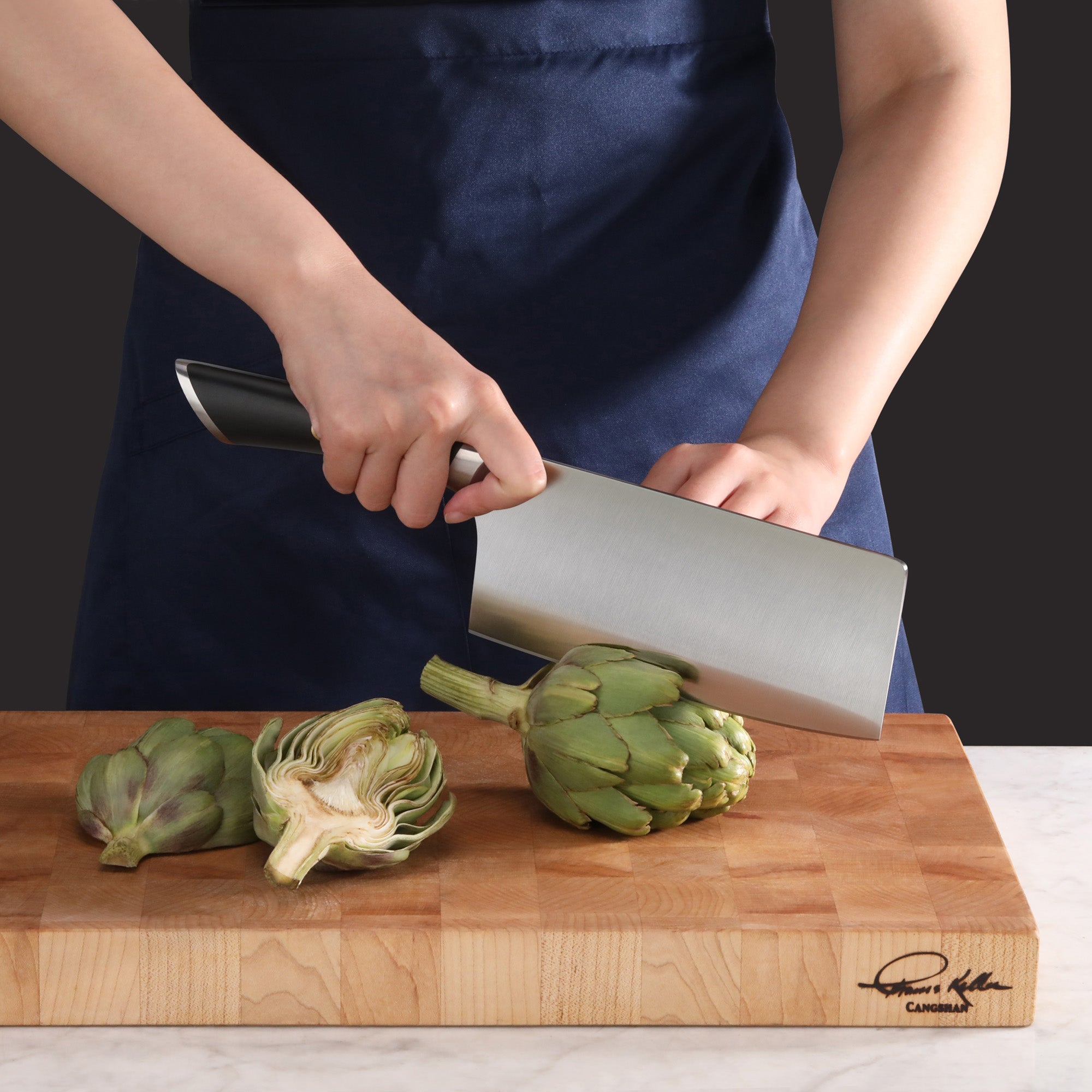 Person wearing a blue apron cutting an artichoke on a wooden cutting board with a large cleaver knife.