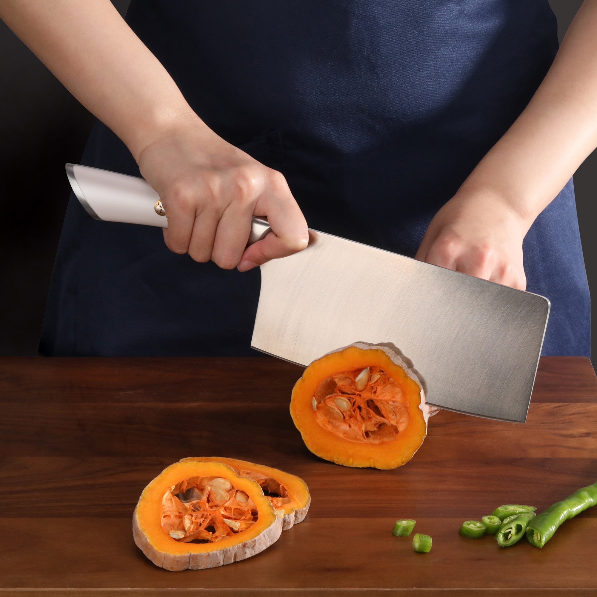 Hand holding a cleaver knife cutting an orange pumpkin on a wooden surface with sliced green chili peppers nearby.