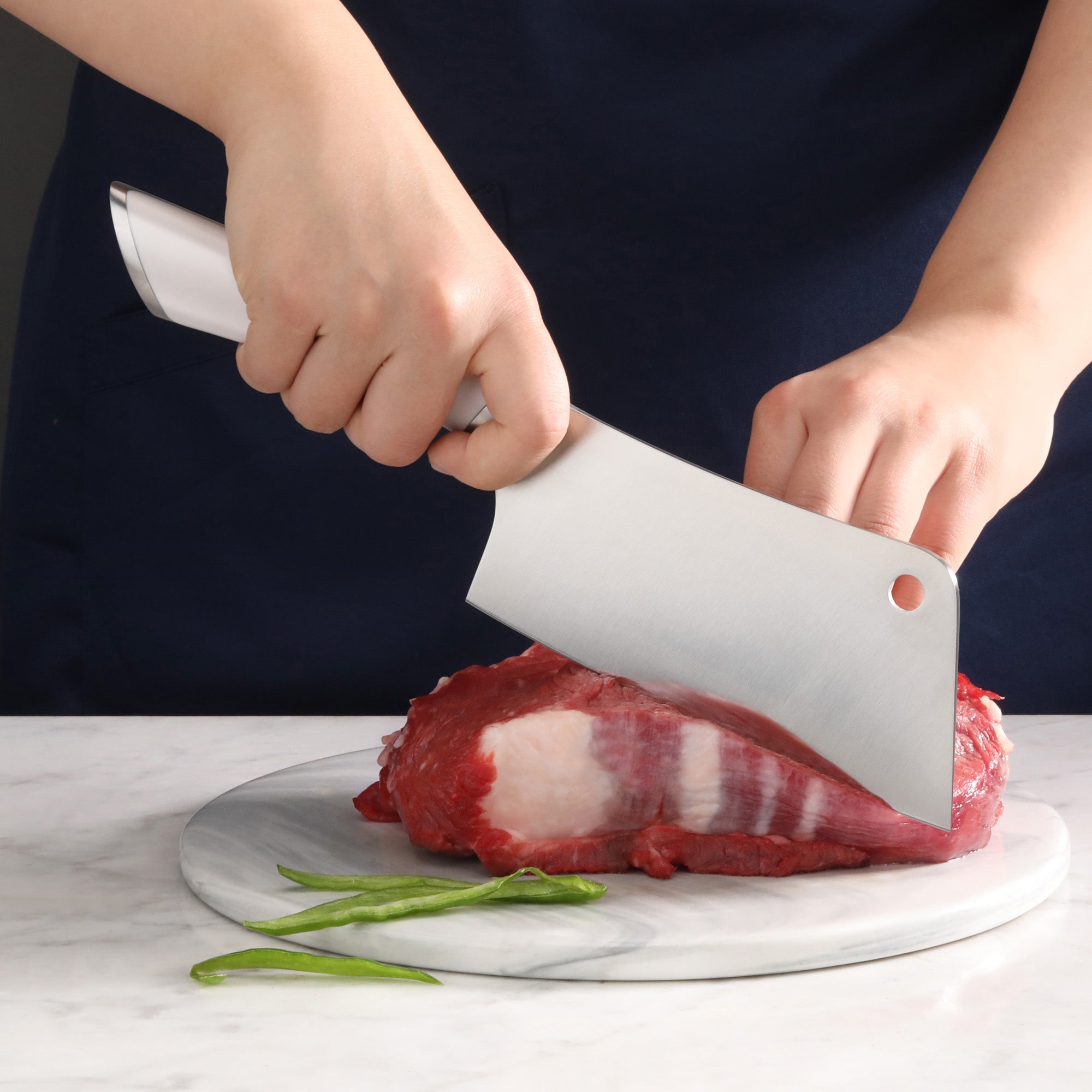 Hands holding a cleaver knife cutting raw beef on a round marble cutting board with green chili peppers nearby.