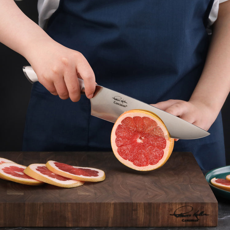 Person holding a chef's knife cutting a grapefruit on a wooden cutting board with sliced grapefruit pieces nearby.