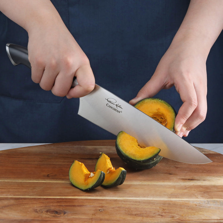 Person slicing a green and orange squash on a wooden cutting board with a large chef's knife.