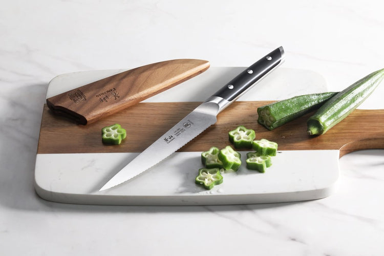 Serrated utility knife with black handle on a cutting board next to sliced and whole okra, with wooden sheath nearby.