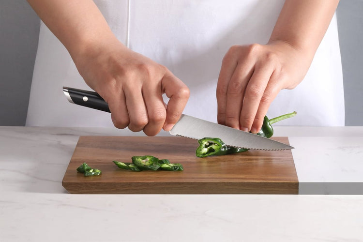 Hands slicing a green chili pepper on a wooden cutting board using a serrated utility knife.