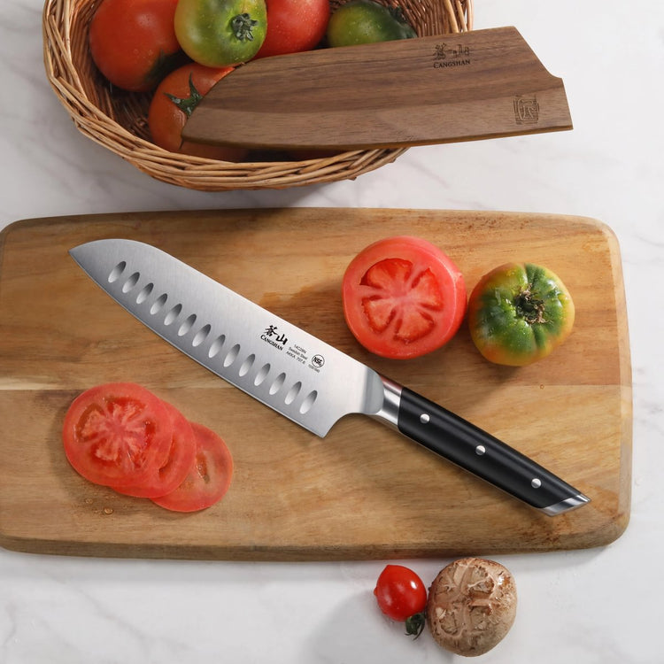 Santoku knife with black handle on wooden cutting board next to sliced and whole tomatoes, basket, and mushroom.