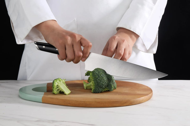 Hands holding a chef's knife cutting broccoli on a wooden cutting board with a green handle.