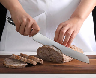 
                    
                      Person slicing a loaf of bread on a wooden cutting board using a serrated knife with a black handle.
                    
                  