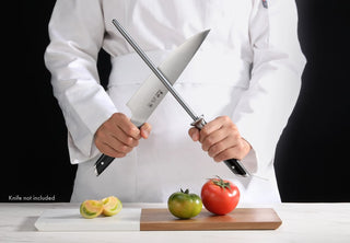
                    
                      Person in white chef coat holding a honing steel and a knife above a cutting board with a green and a red tomato and sliced l
                    
                  