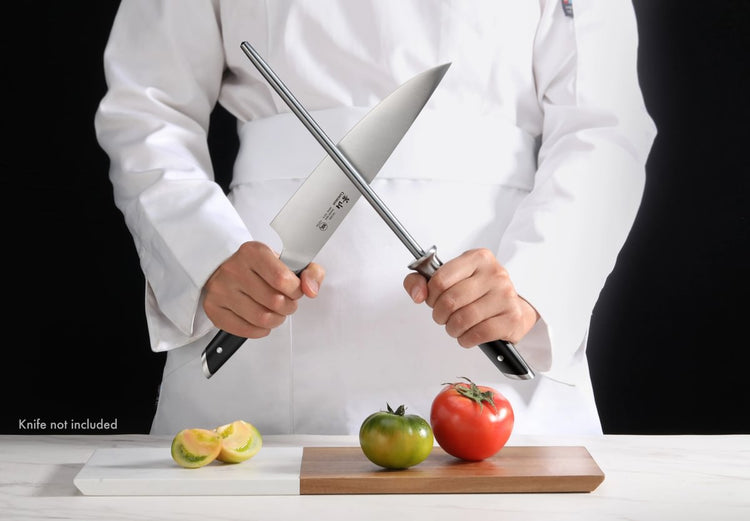 Person in white chef coat holding a honing steel and a knife above a cutting board with a green and a red tomato and sliced l