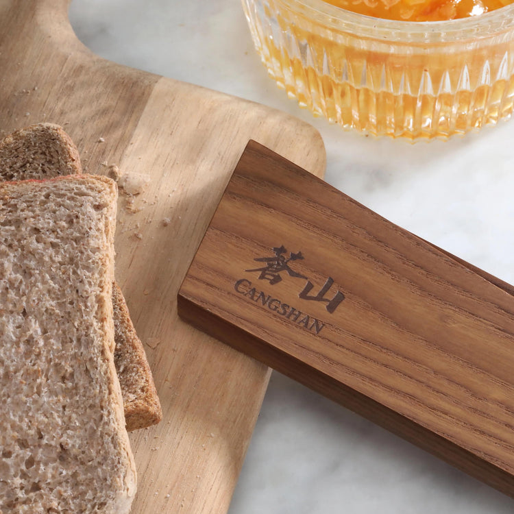 Ash wood magnetic knife sheath with engraved logo next to sliced whole wheat bread on wooden cutting board.