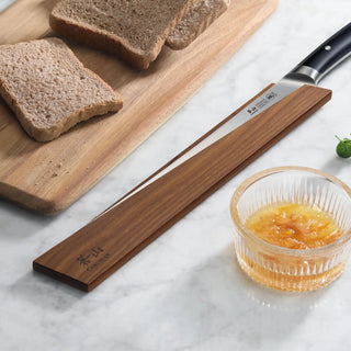 
                    
                      Ash wood magnetic knife sheath covering a slicing knife blade on a marble surface next to bread and a small glass bowl of jam
                    
                  