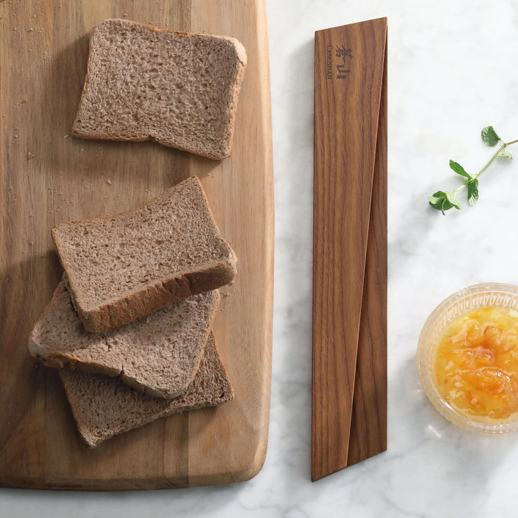 Ash wood magnetic knife sheath placed next to sliced bread on a wooden cutting board and a small bowl of orange jam.
