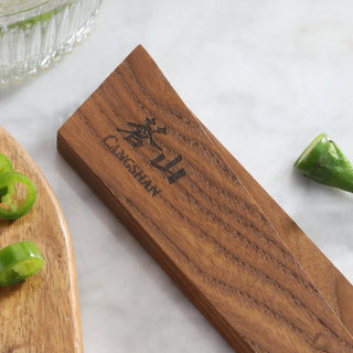 
                    
                      Ash wood magnetic knife sheath with engraved logo, placed on a marble surface near sliced green peppers.
                    
                  
