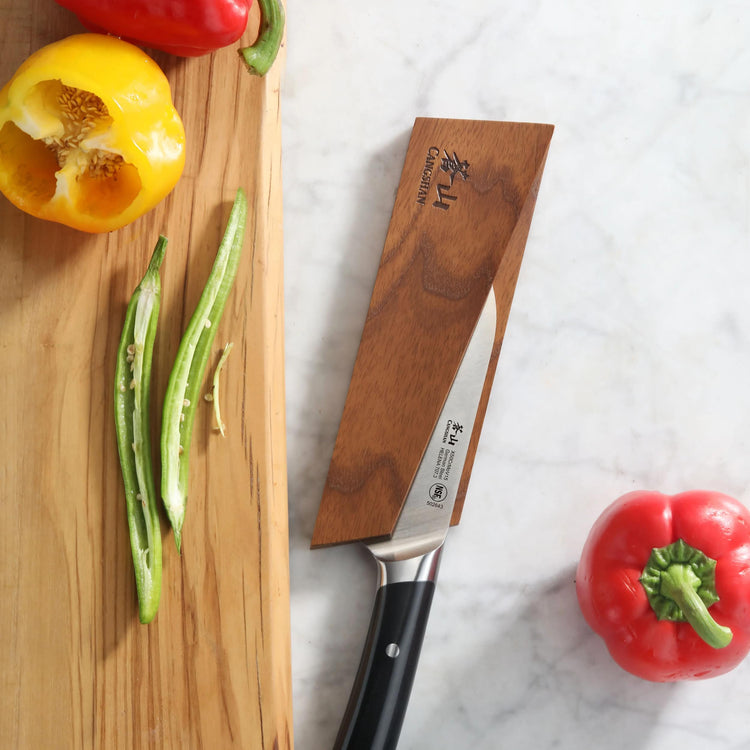 Ash wood magnetic knife sheath covering a kitchen knife blade on a marble surface next to sliced vegetables.