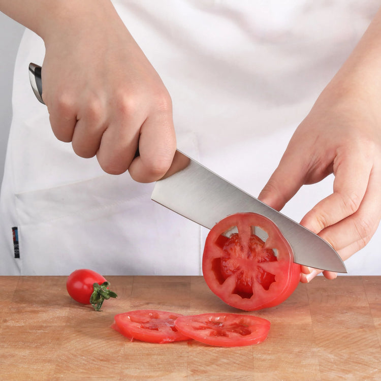 Hand holding a knife slicing a tomato on a wooden cutting board with two tomato slices and a cherry tomato nearby.