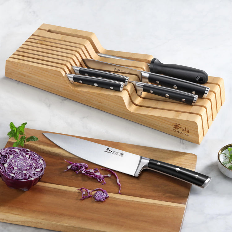 Seven knives with black handles stored in a wooden in-drawer organizer, with one chef knife slicing red cabbage on a cutting