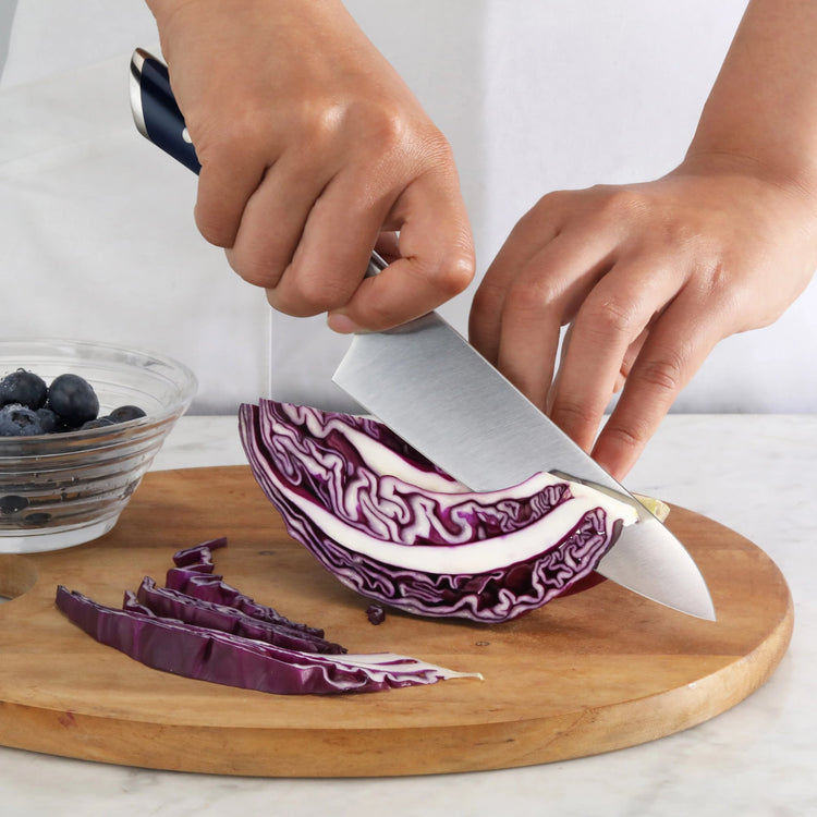 Hands slicing purple cabbage on a wooden cutting board with a navy and white-handled knife.