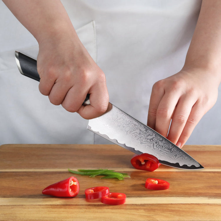 Hand holding a chef's knife slicing a red bell pepper on a wooden cutting board.