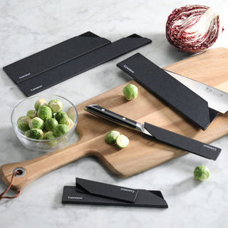 
                    
                      Six black knife edge guards of varying sizes on a wooden cutting board and marble surface with a knife, Brussels sprouts, and
                    
                  