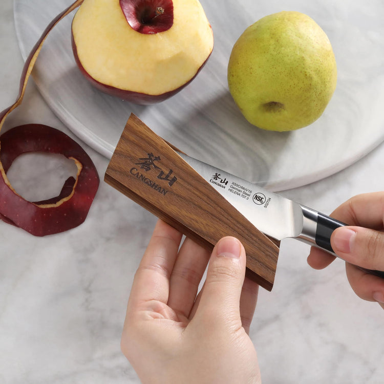 Hand holding a small knife with a wooden magnetic sheath near a peeled apple and a pear on a marble surface.