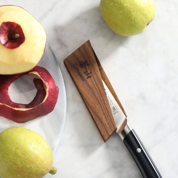 Ash wood magnetic knife sheath holding a paring knife next to peeled and whole apples on a marble surface.