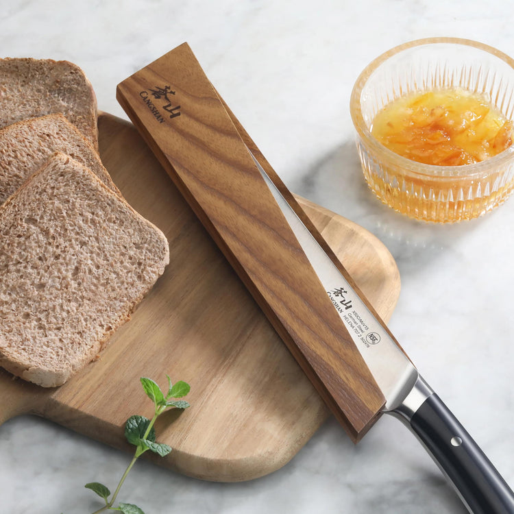 Ash wood magnetic knife sheath covering a bread knife on a wooden cutting board with sliced bread and a glass bowl of marmala