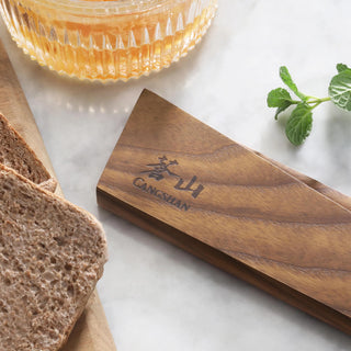 
                    
                      Ash wood magnetic knife sheath with engraved logo placed on a marble surface next to sliced bread and a glass bowl.
                    
                  